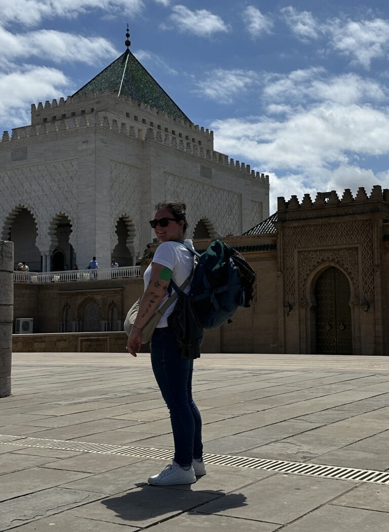 Solo female traveller with underseat backpack standing in the courtyard of the Mausoleum of Mohammed V in Rabat, Morocco. with its ornate arches and green-tiled tower roof in the background.