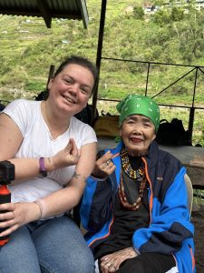 A solo female traveller makes heart shapes with her hands with tattoo artist Apo Whang Od doing the same.
